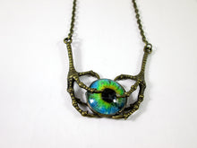 Load image into Gallery viewer, A gothic-style necklace featuring a detailed, claw-like bronze metal frame holding a round cabochon glass eye pendant with a vivid blue, green, and black iris.