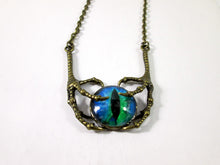 Load image into Gallery viewer, A gothic-style necklace featuring a central glass cabochon eye with blue and green iridescence and a black slit pupil, held in an antique bronze claw-like frame with a matching chain.