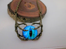 Load image into Gallery viewer, A fantasy-style necklace featuring a glowing blue and teal eye pendant with a black slit pupil, suspended in an intricate, textured bronze claws, displayed on a rustic wood slice stand against a plain white background.
