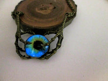Load image into Gallery viewer, A handmade pendant featuring a blue and green iridescent eye-shaped bead suspended in a pair of antique bronze claws, displayed on a rustic wood slice against a plain light background.