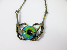 Load image into Gallery viewer, A gothic-style necklace featuring a central glass cabochon eye pendant with a vivid blue, green, and black iris, held by two textured, clawed metal hands in an antique bronze finish, suspended from a matching chain.