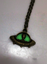 Load image into Gallery viewer, An antique bronze UFO pendant necklace featuring a green glowing insert with a bronze alien silhouette and the engraved text 'I WANT TO BELIEVE', hanging from a bronze chain on a light background.