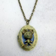 Load image into Gallery viewer, An oval locket pendant necklace featuring a small mouse figure in front of a blue background, all in an bronze finish, hanging from a matching chain against a light textured backdrop.