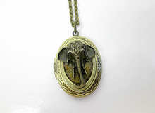 Load image into Gallery viewer, An oval locket pendant necklace featuring a detailed elephant head design in antiqued bronze, suspended from a matching chain against a plain white background.
