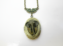 Load image into Gallery viewer, An oval locket pendant necklace featuring a detailed elephant head design in antiqued bronze, suspended from a triangular filigree connector and matching chain against a plain white background.