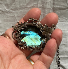 Load image into Gallery viewer, A close-up of a hand holding an oval pendant featuring a shimmering blue-green labradorite stone set in a copper frame with a butterfly and vine detail, against a wrinkled light gray fabric background.  