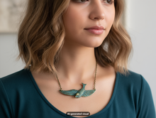 Load image into Gallery viewer, A close-up of a woman wearing a antique bronze sparrow necklace with verdigris patina, paired with a dark teal top. The image has an 'AI-generated visual' watermark at the bottom.