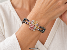 Load image into Gallery viewer, Close-up of a pink flower magnetic hematite bracelet featuring a floral centerpiece with pink and gold accents on a beaded band.  A small "AI-generated visual" label appears at the bottom of the image.