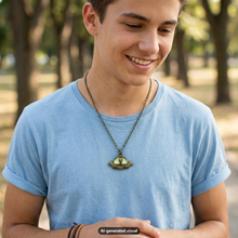 Load image into Gallery viewer, A young man wearing a light blue t-shirt and a bronze-toned lit-up green UFO alien necklace, standing in a park with blurred trees in the background.  A small "AI-generated visual" label appears at the bottom.