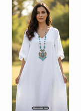 Load image into Gallery viewer, A woman wearing a loose white V-neck kaftan dress with elbow-length sleeves, styled with a long turquoise-and-silver pendant with turquoise beaded tassel necklace, standing outdoors against a blurred green background. A small label at the bottom reads 'AI-generated visual'.