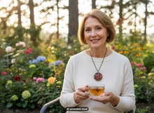 Load image into Gallery viewer, A smiling older woman sits in a garden holding a glass cup of tea. She wears a cream knit sweater and a vintage red hummingbird locket necklace.  A small "AI-generated visual" label appears at the bottom.