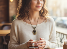 Load image into Gallery viewer, A woman in a cozy cafe setting wearing a cream knit sweater and a bronze-toned elephant locket necklace, holding a white mug. The image has a warm, soft-focus aesthetic with a visible 'AI-generated visual' watermark at the bottom.